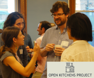 Photo of four adults standing in circle in a room, holding drinks and talking.