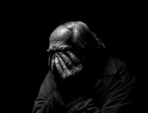 Black and white photo with black background. Foreground is person shown shoulders up, covering their face with their hands.