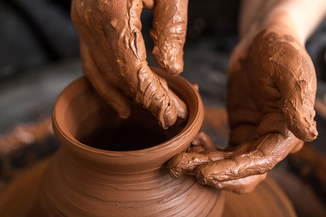 Photo of clay covered hands making a small pot on a potter's wheel.