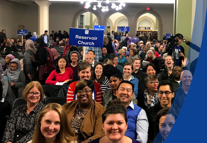 Image of people's faces seated in a large room, smiling and looking at camera. One person holds sign that reads "Reservoir Church."