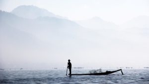 Photo of fisherman silhouette, standing on small boat in water. Mist and mountains in background.
