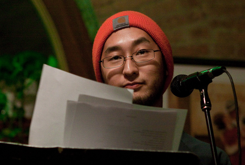 Asian male with glasses, orange knit cap holding papers at microphone.